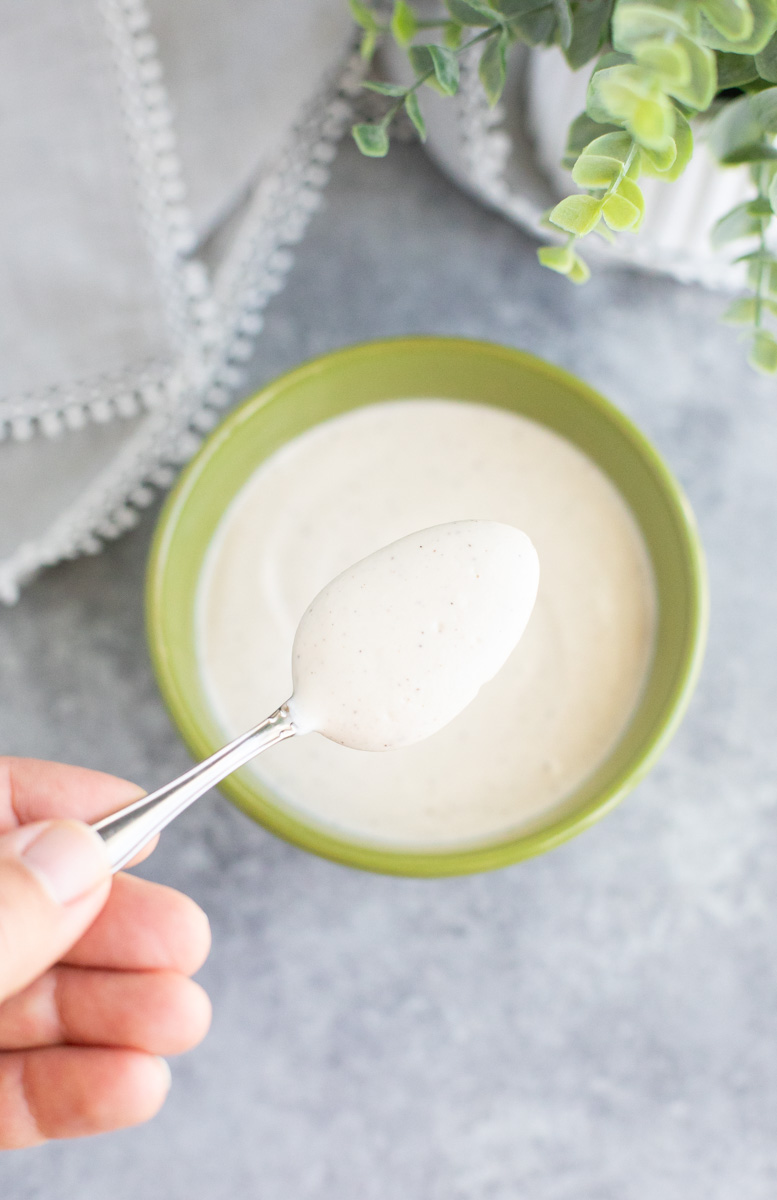 A hand holding a spoonful of creamy white sauce over a green bowl on a gray background.