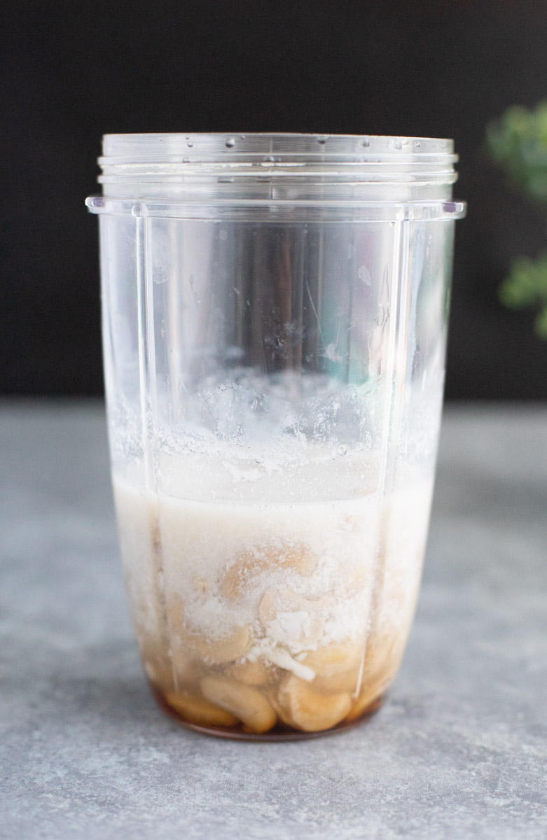 A clear blender cup filled with liquid and cashews on a gray background.
