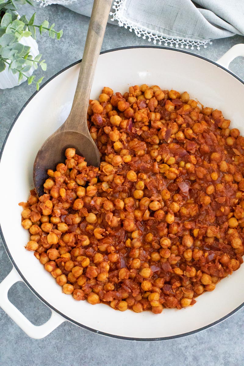 A white pan filled with chickpeas in red sauce with a wooden spoon on a gray background. 