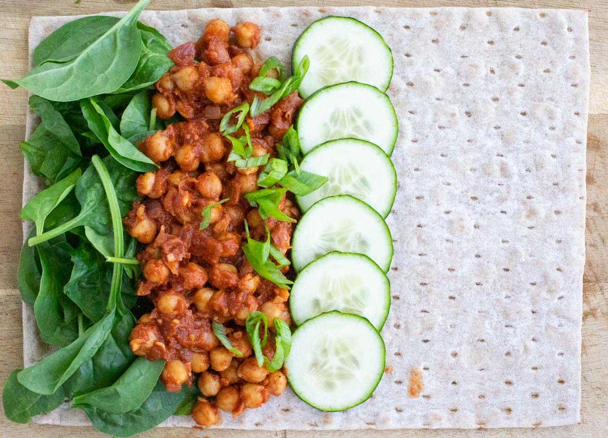 A lavash wrap topped with spinach, chickpeas, and cucumbers on top of a wooden cutting board. 