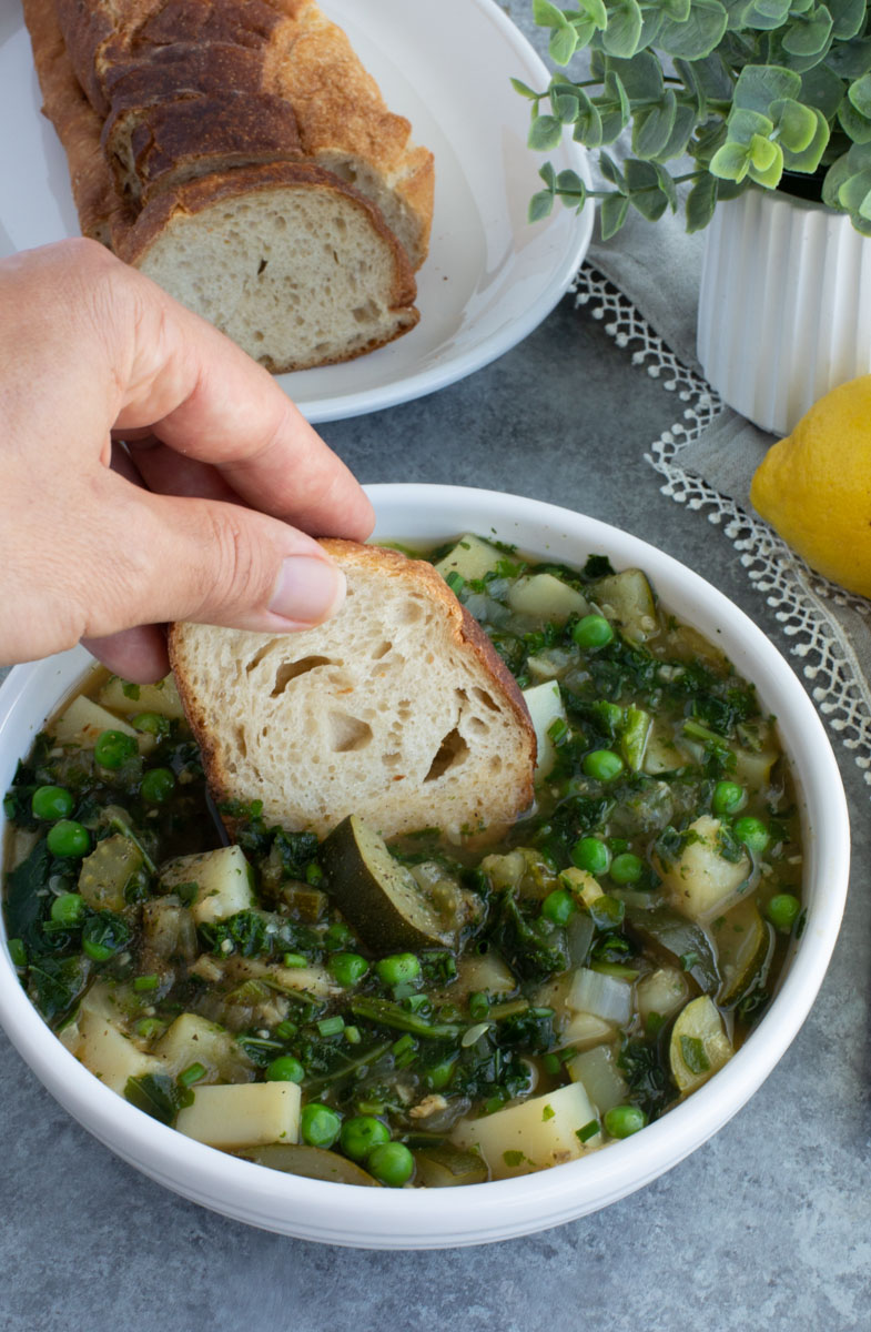 A hand dipping a piece of bread into a white bowl filled with soup on a gray background.