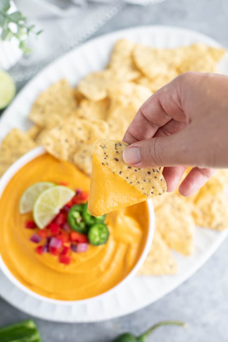 A hand dipping a tortilla chip into a bowl of vegan cheese next to a platter filled with chips on a gray background.