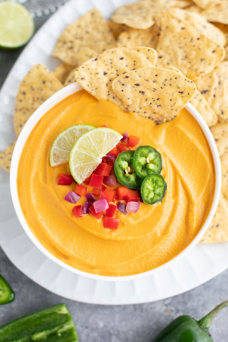 A white bowl filled with an orange dip, toppings, and two tortilla chips on top of a platter filled with chips. 