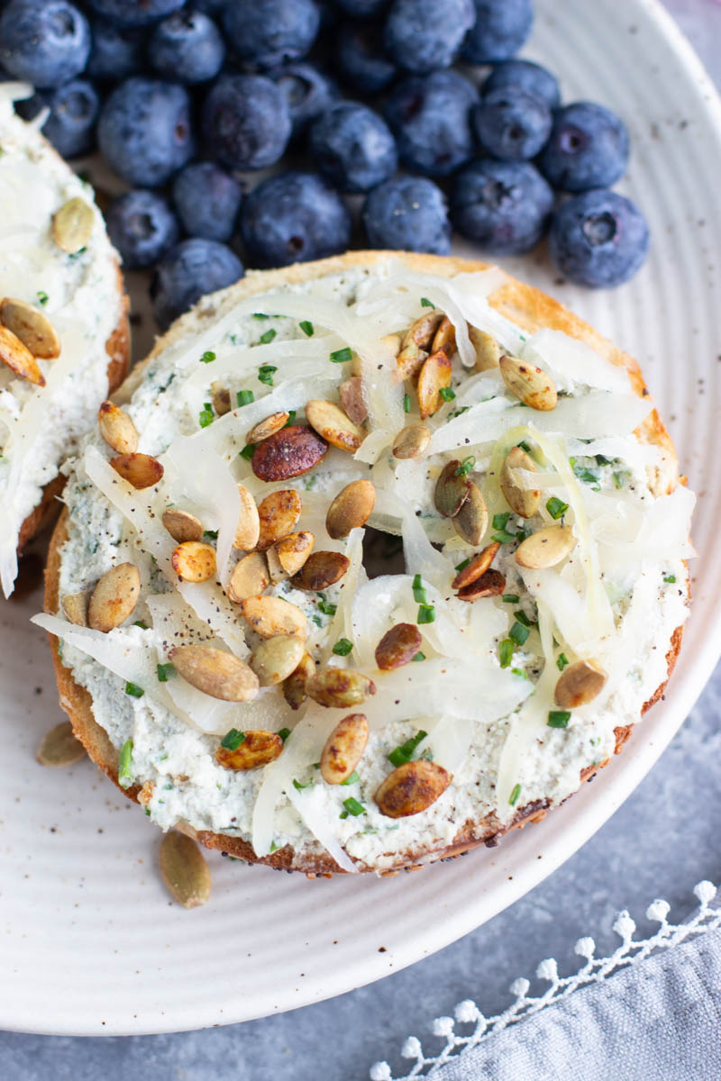 Half a bagel topped with vegan cream cheese, pumpkin seeds, sauerkraut, and chives next to blueberries on a white plate.