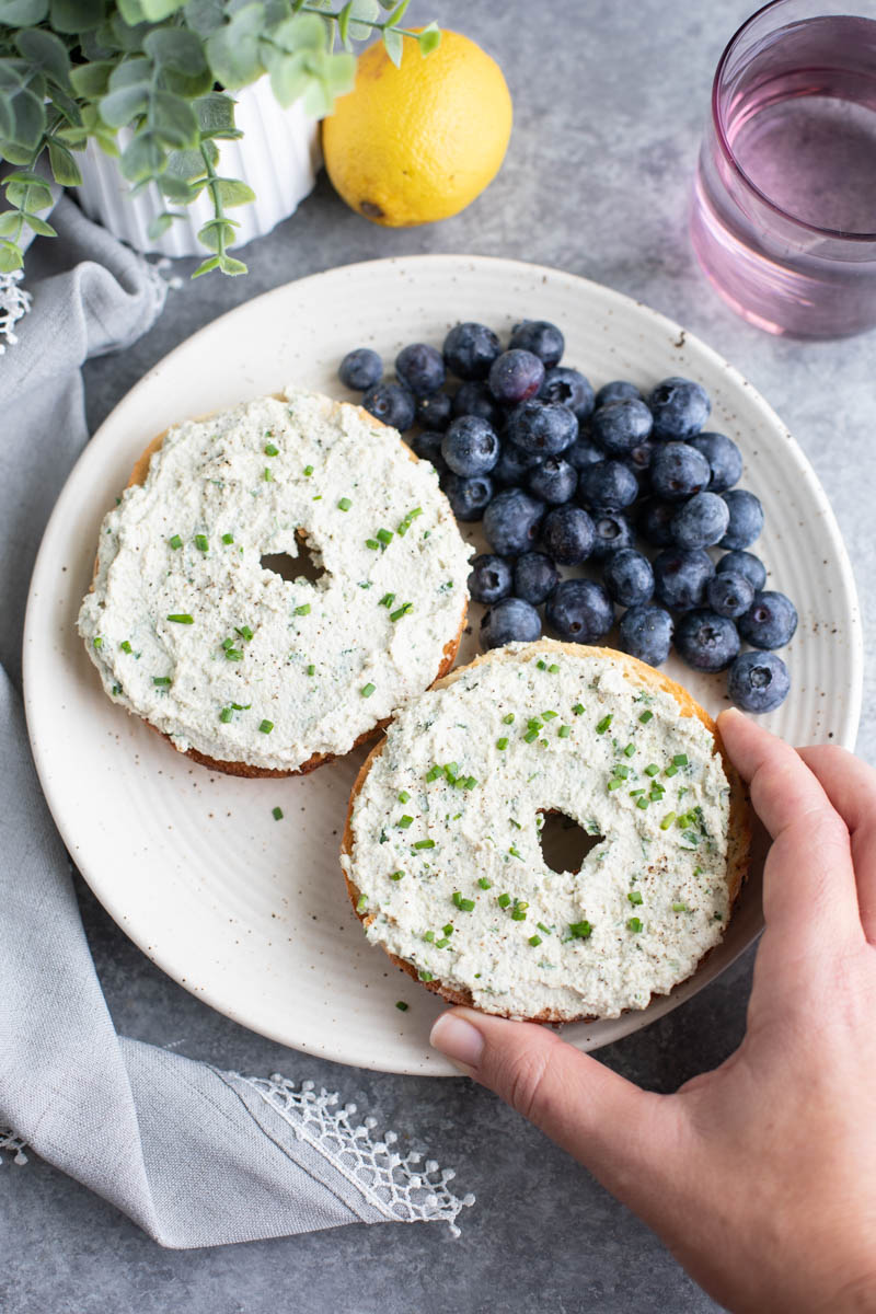 A hand grabbing half a bagel from a plate with another half and blueberries on a grey background. 