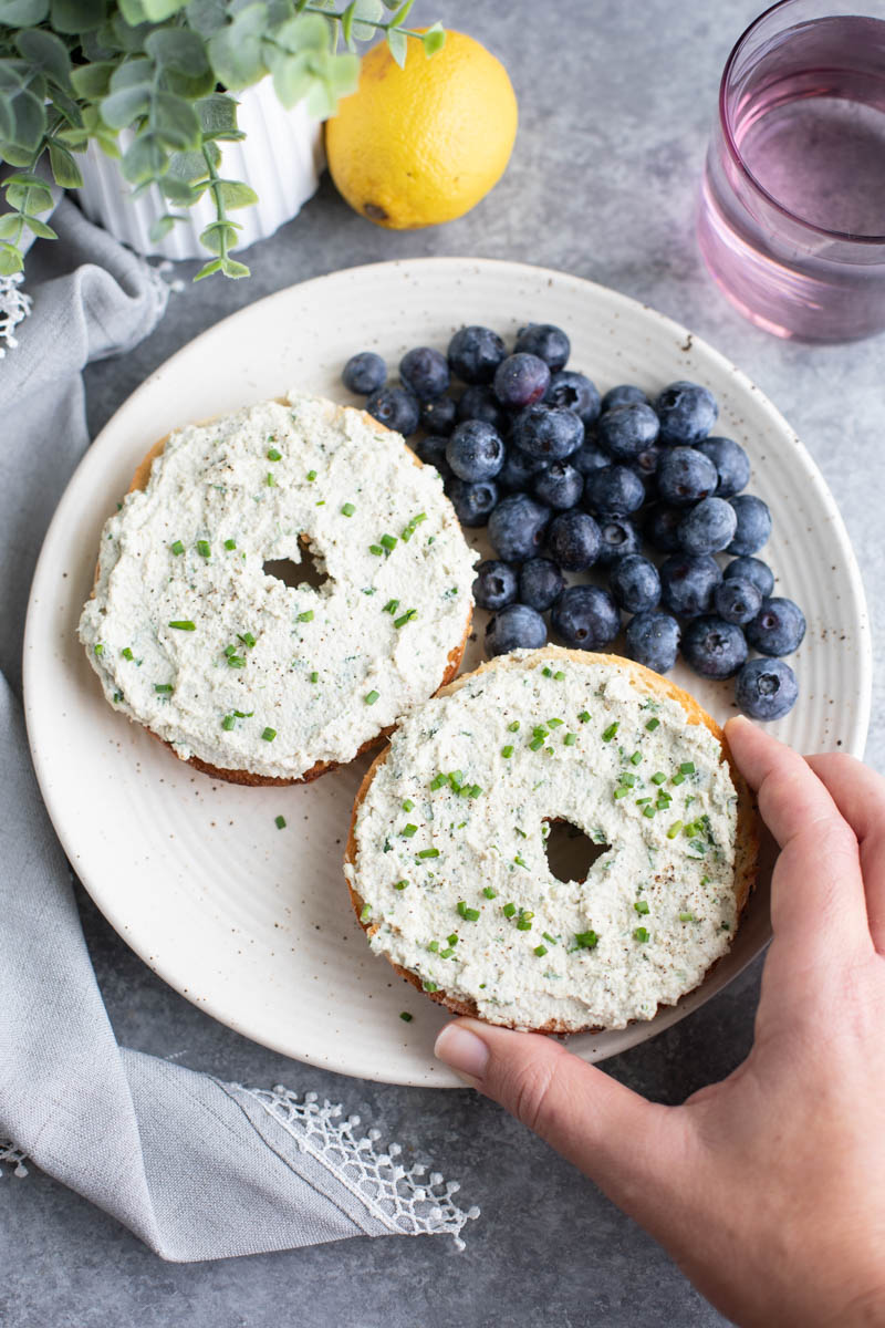 A hand grabbing half a bagel from a plate with another half and blueberries on a grey background.