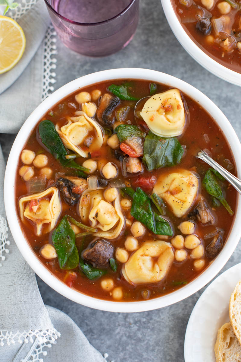 A white bowl filled with vegan tortellini, vegetables, and chickpeas and a spoon on a gray background. 