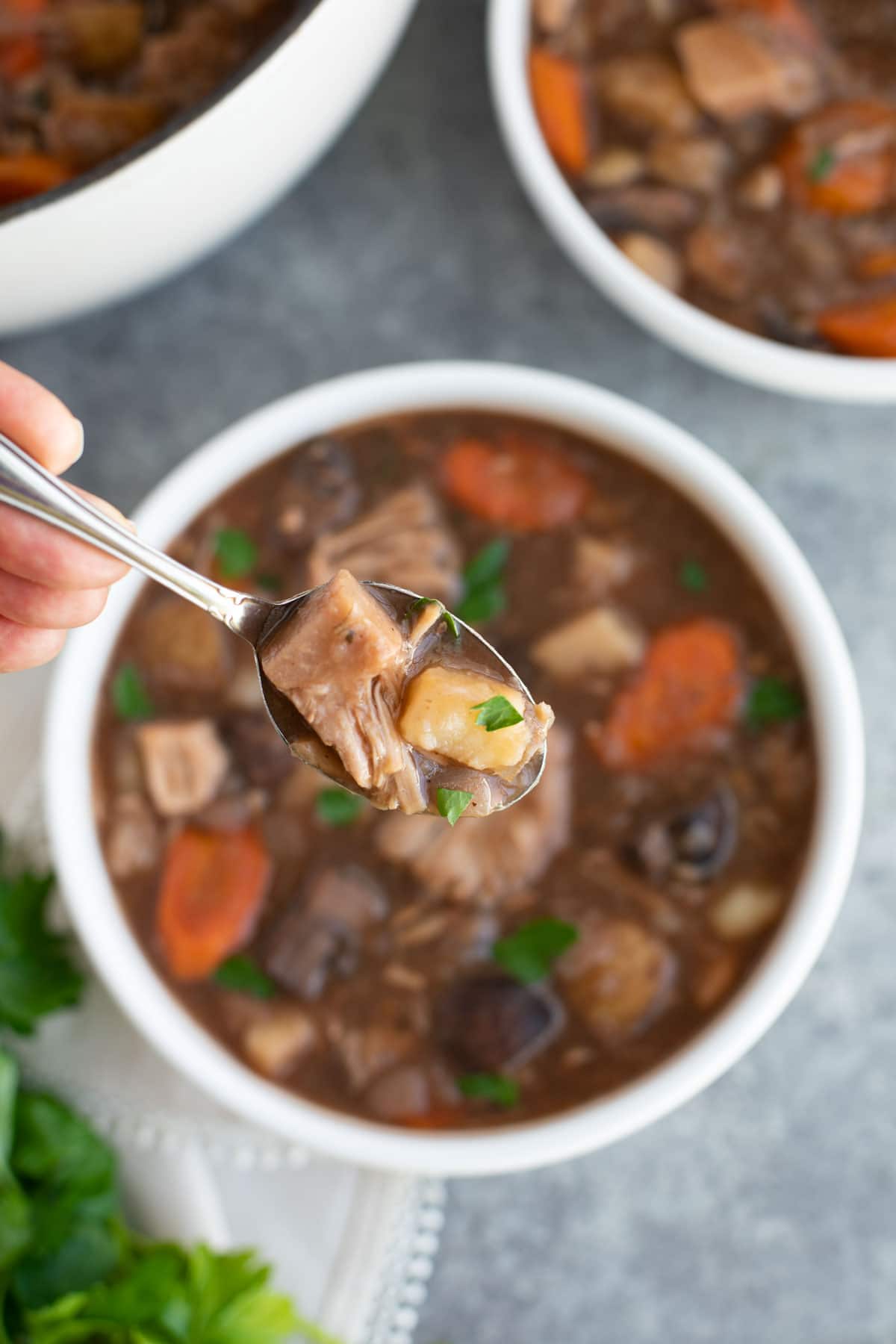 A hand holding a spoonful of stew over a bowl on a gray background.