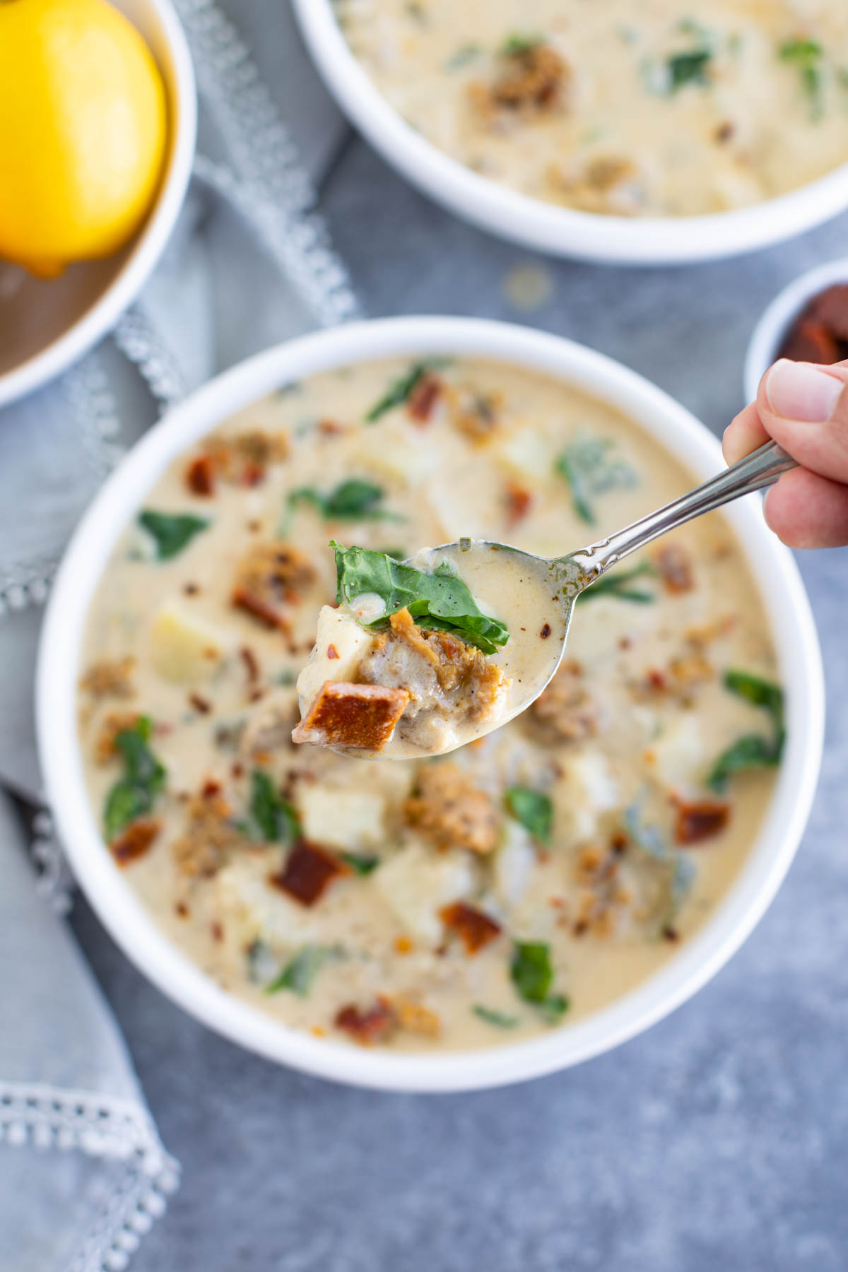 A hand holding a spoon over a white bowl filled with soup on a gray background.